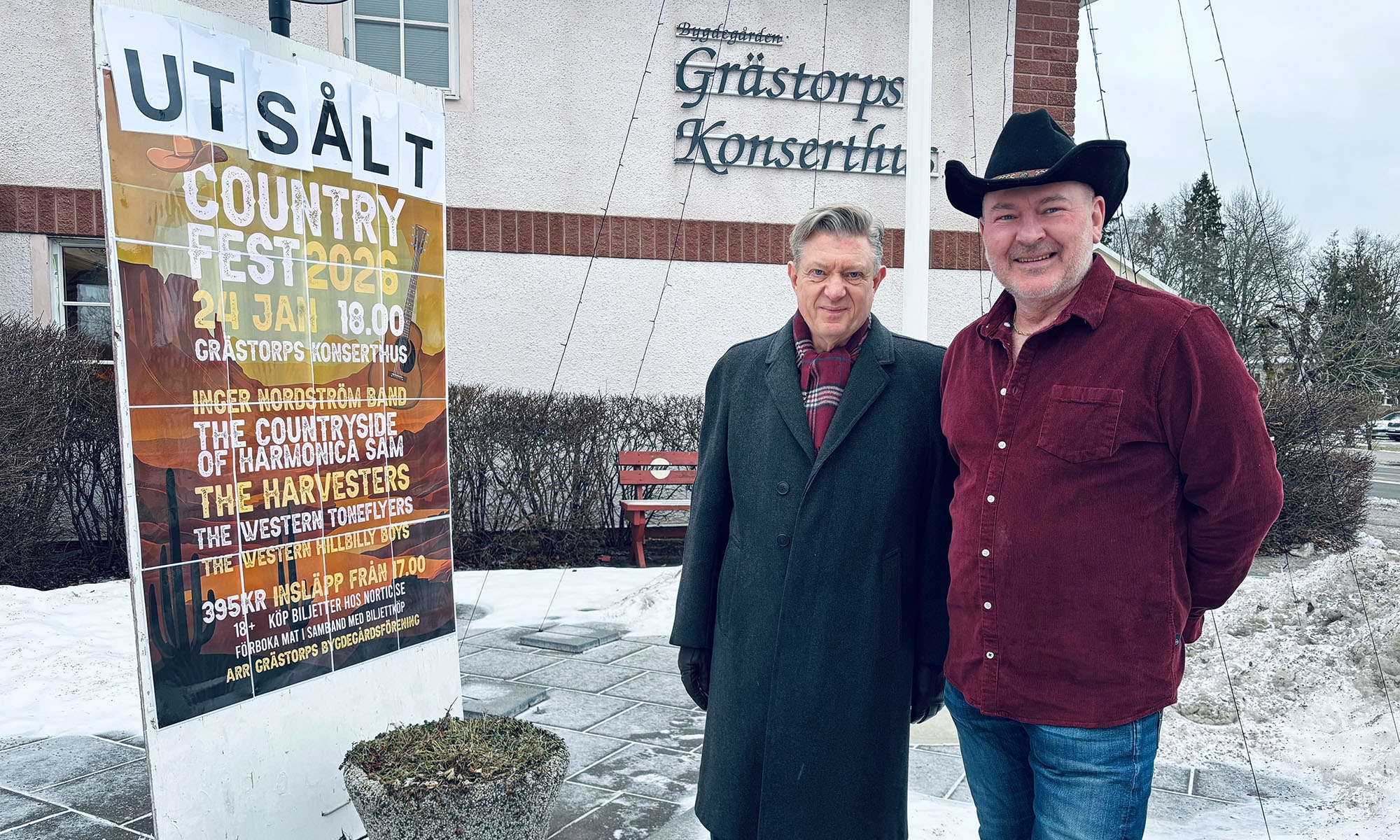 Två män som står framför ett hus. En har på sig en hatt.